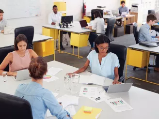 Small workgroup meeting with printed documents on the desk and office workers at desks in the background